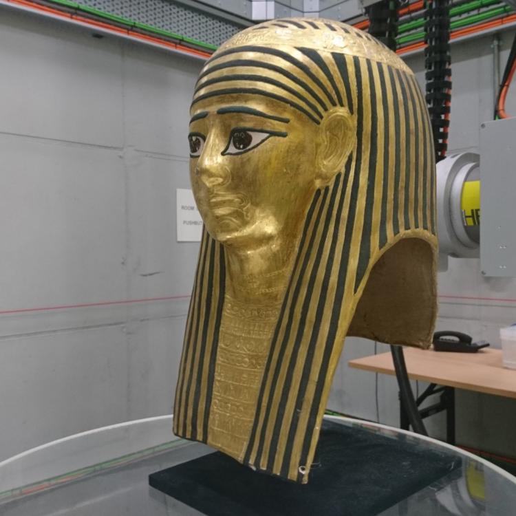 Gold bust of an Egyptian figure wearing a nemes headdress, resting on a large cylindrical stand in a room with mounted scanning equipment and cable rigging in the background.