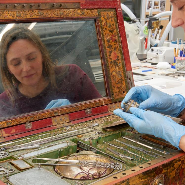 A conservator assesing a weight taken from an open painted wooden box with a mirror inside. 