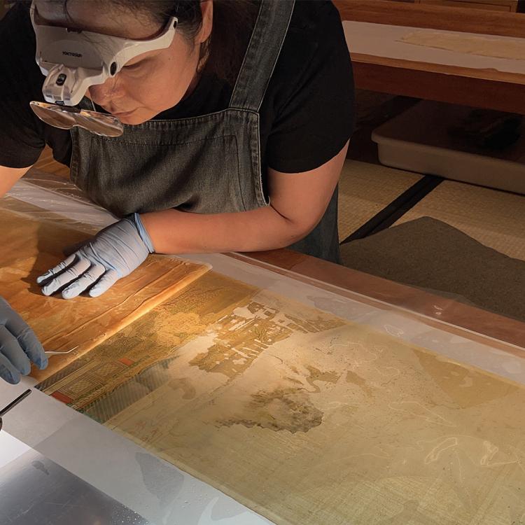 A conservator wearing magnifying glasses and gloves working on a screen painting.