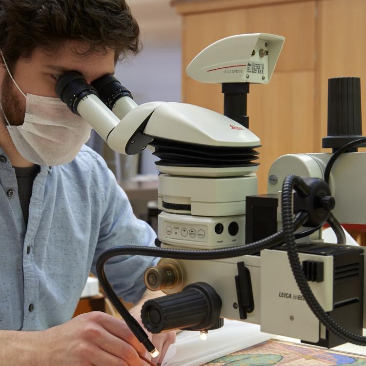 Conservator seated at a desk looking through a microscope while working on a painting