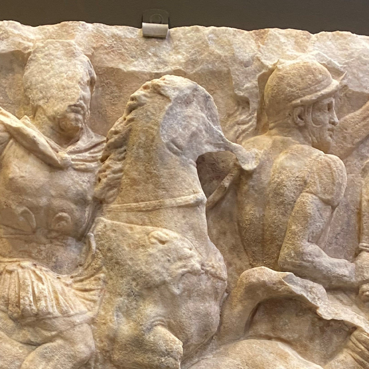 A stone relief of a man with a cloak riding a horse which tramples a person beneath it, and another soldier holding a sword.