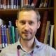 A photograph of Dr Richard Hobbs in a grey shirt, standing in front of a bookshelf.