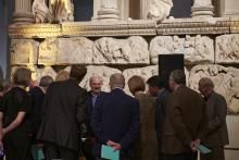 A group stood in a gallery, some with pamphlets held behind their back, listening to a speaker in front of the Nereid Monument