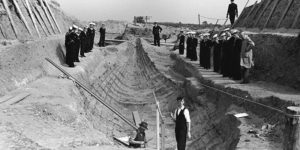 Photograph of two people standing in the ship excavation at Sutton Hoo with at least 20 sailors standing above. Everyone looking towards the camera.