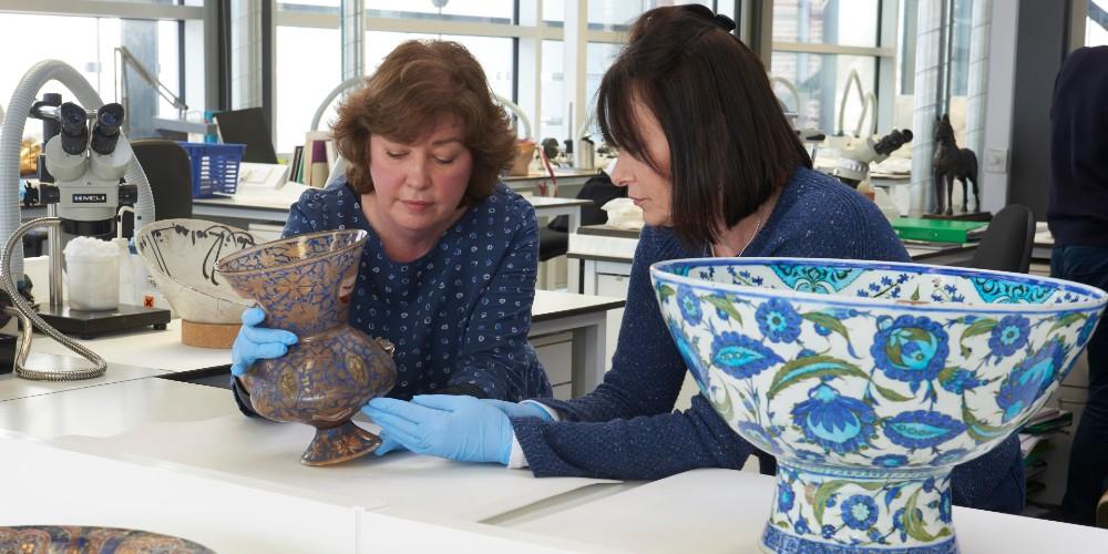 Two conservators looking closely at a lamp from a mosque with a painted ceramic object in the foreground. 