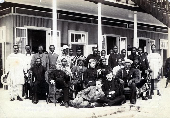 Portrait of a group of men seated and standing in front of a colonial style building.