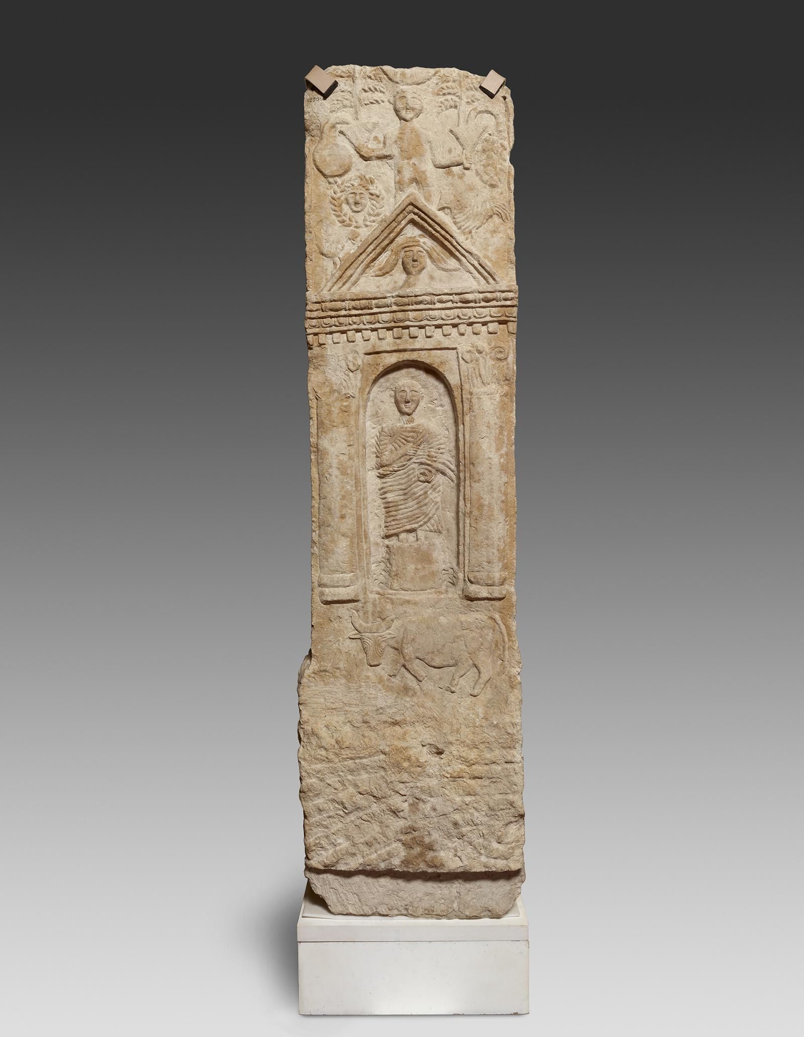 Limestone stela, gable missing, surface worn in places. A female figure stands on a high pedestal in a round-arched niche within a gabled shrine.