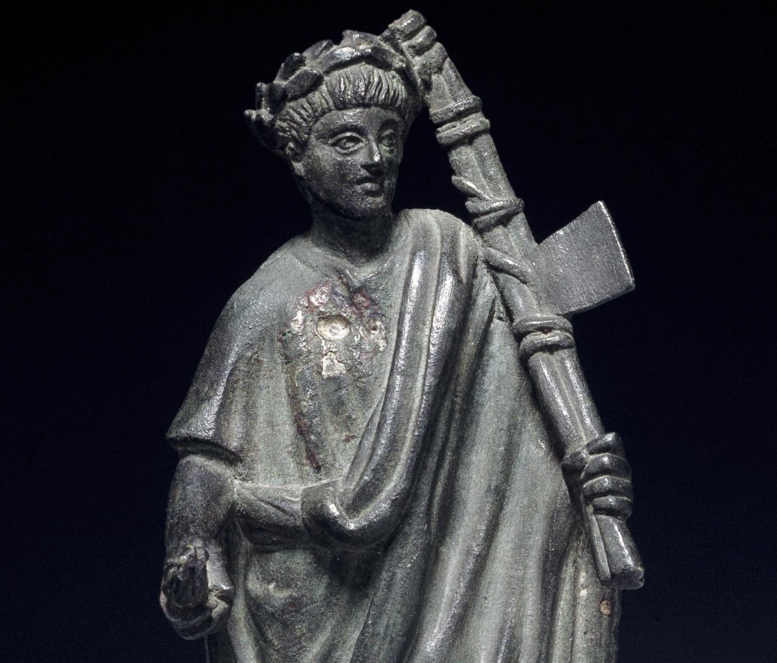 Bronze figure of a lictor (magistrate's attendant) wearing a toga and a wreath on his head. He carries laurel leaves in his right hand and in his left hand he carries the fasces, an axe bound to a bundle of rods.