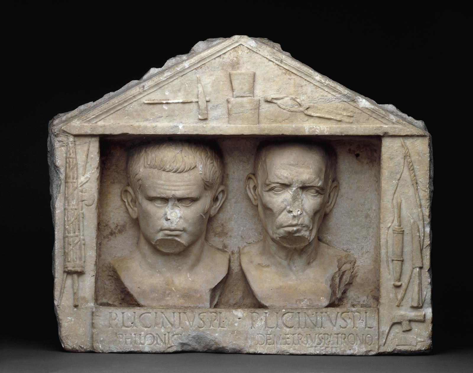 A marble relief with portraits of two human heads. On the left are the rods and axes used in the ceremony of freeing an enslaved individual. In the pediment are the tools of a smith or moneyer, and, on the right, the tools of a carpenter.