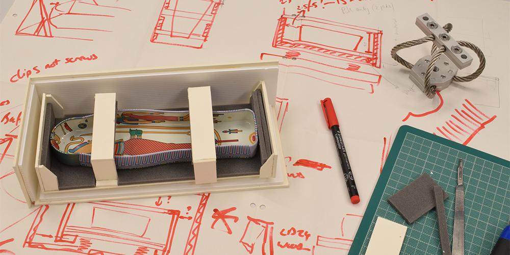 A small model of a mummy in a transport crate, a pen and a steel string on a sheet of paper with sketches of the mummy crate.