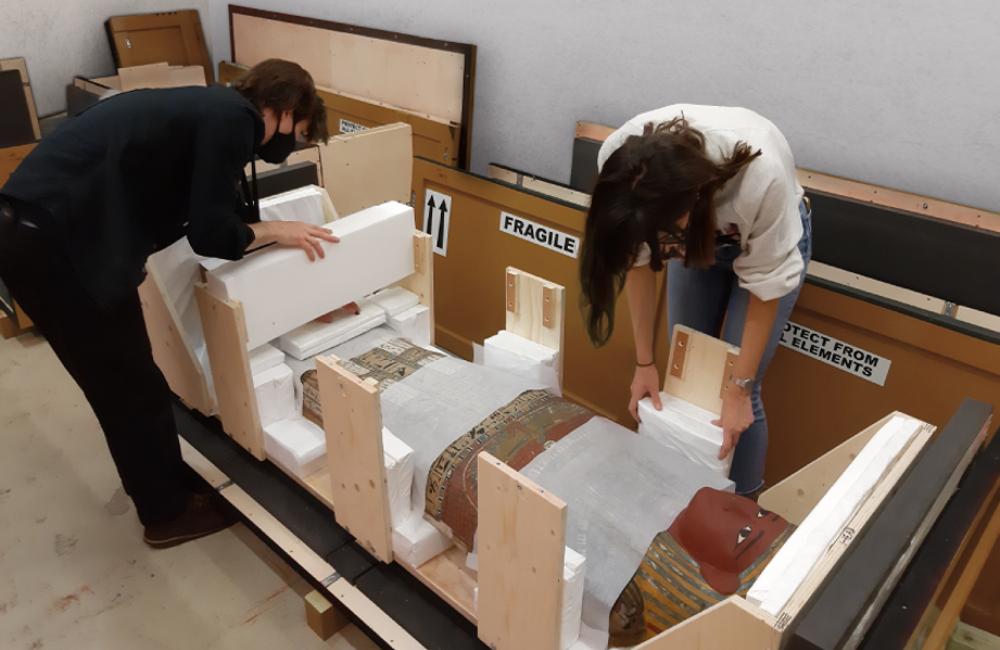 Two people packing the lid of a coffin in a crate. 