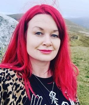 A photo of Laura, who has bright red hair and is wearing a black top with symbols on it, standing in a misty, mountainous landscape.