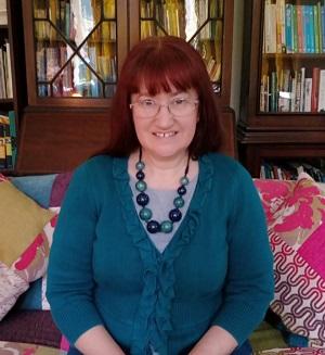 A photo of Lucya, who has red hair and is wearing a blue cardigan, on a colourful sofa,