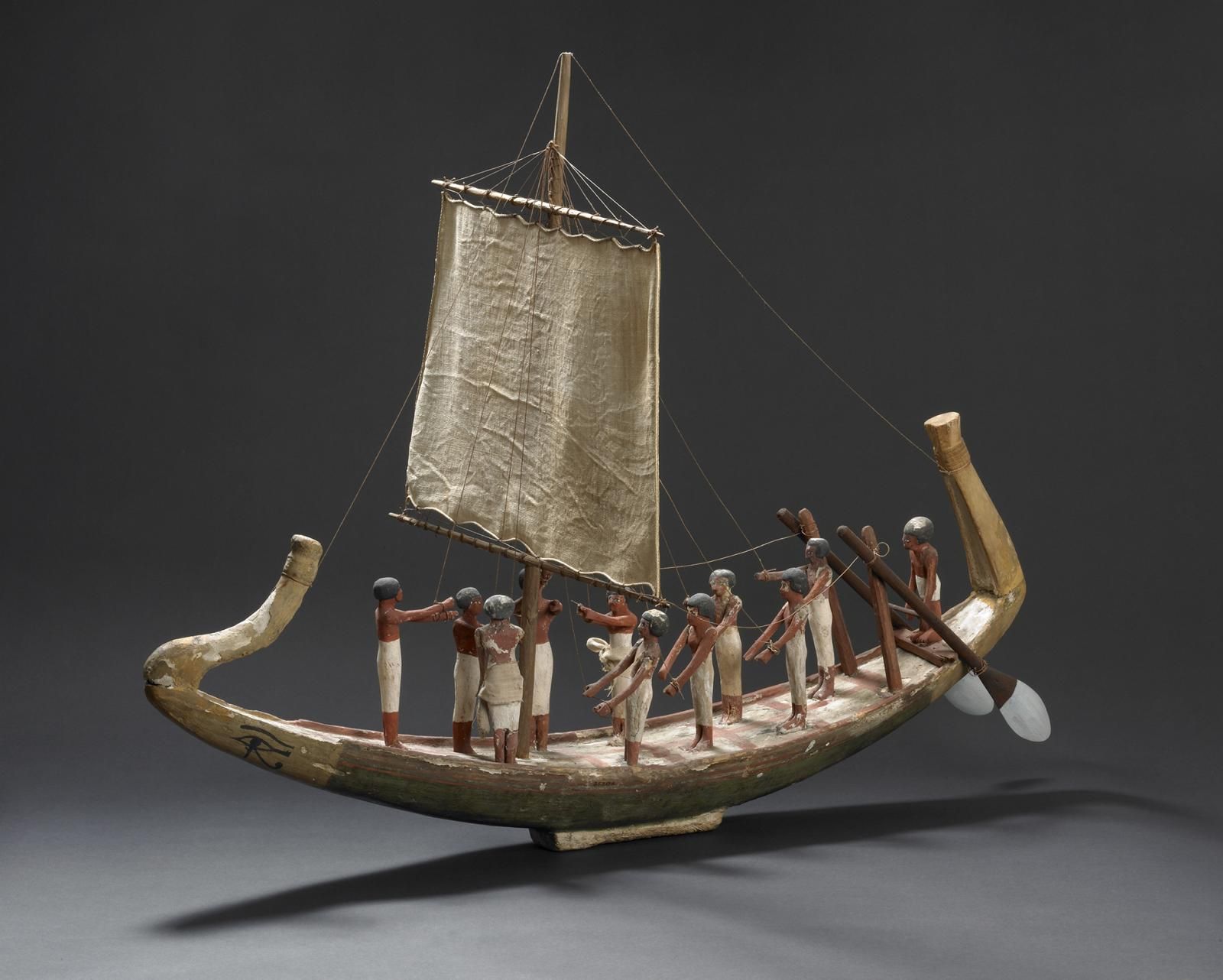 Wooden model of boat with narrow hull and finials each end, sail and small plinth to stand on. Crew consisting of helmsman and ten other hands, all standing