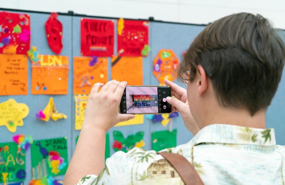 A person viewed from behind, taking a picture of multicoloured messages on a wall. 