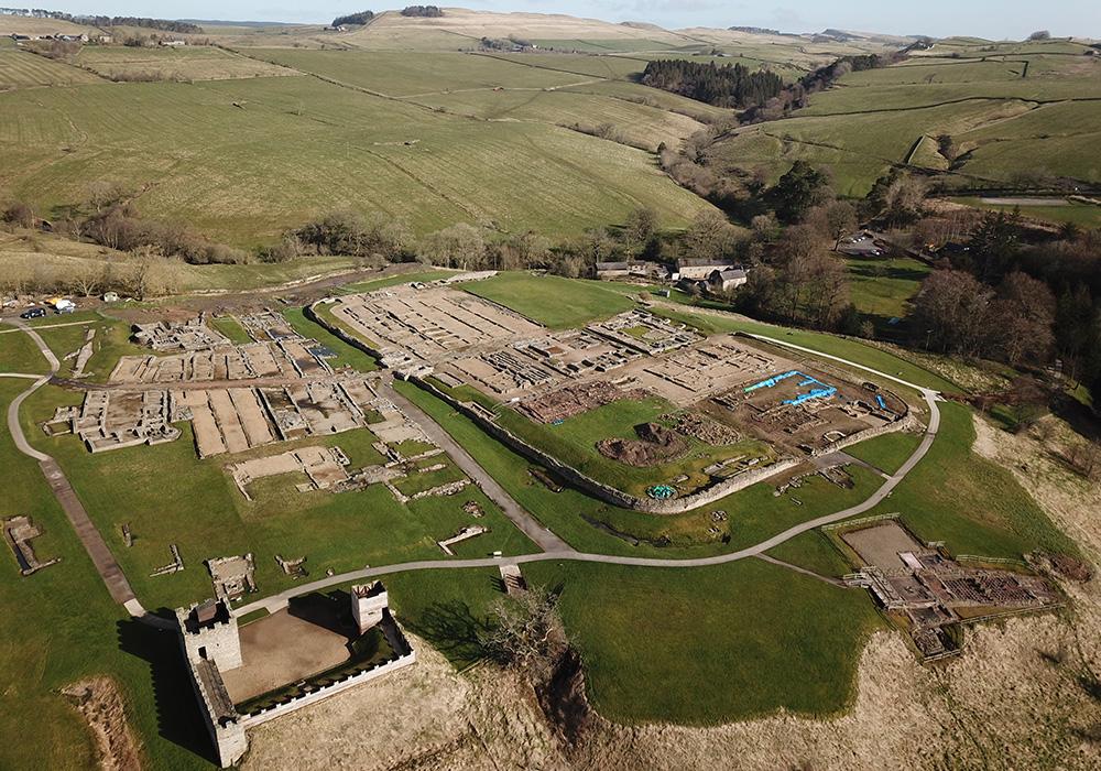 Aerial view of the stone remains of a fort and surrounding fields with a view of the sea in the background. 