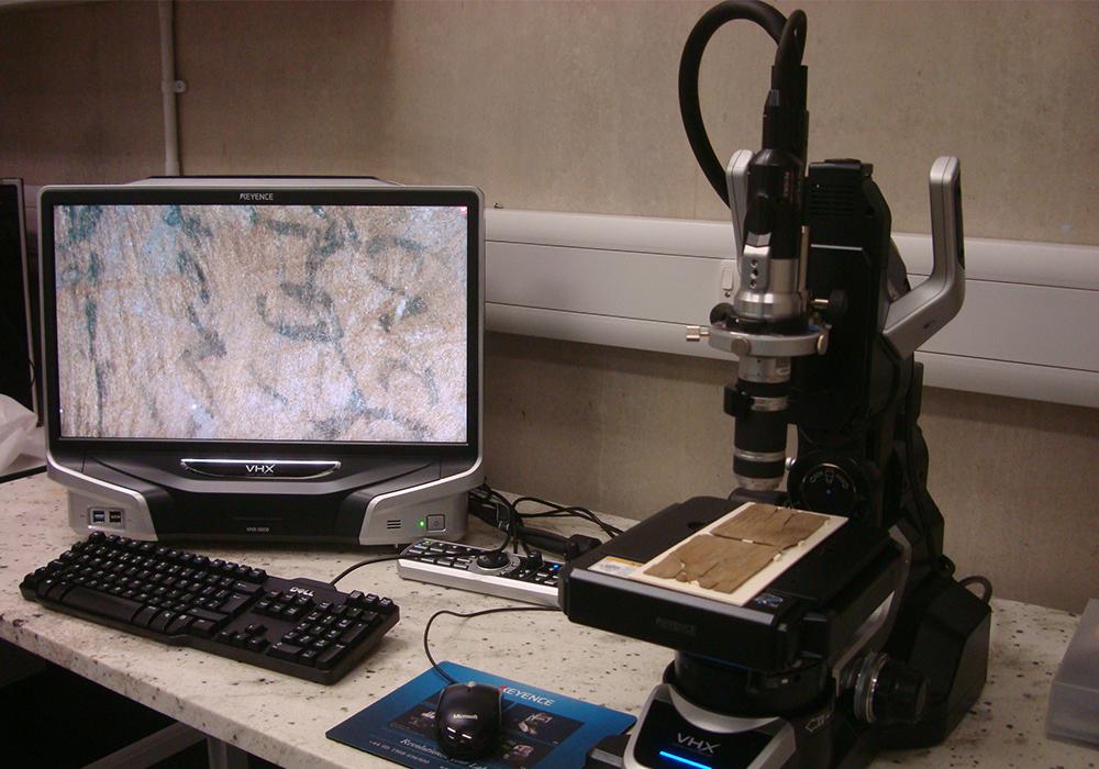 A Vindolanda tablet on a digital microscope. An image on a computer screen shows a close-up of some of the ink writing on the wood tablet. 