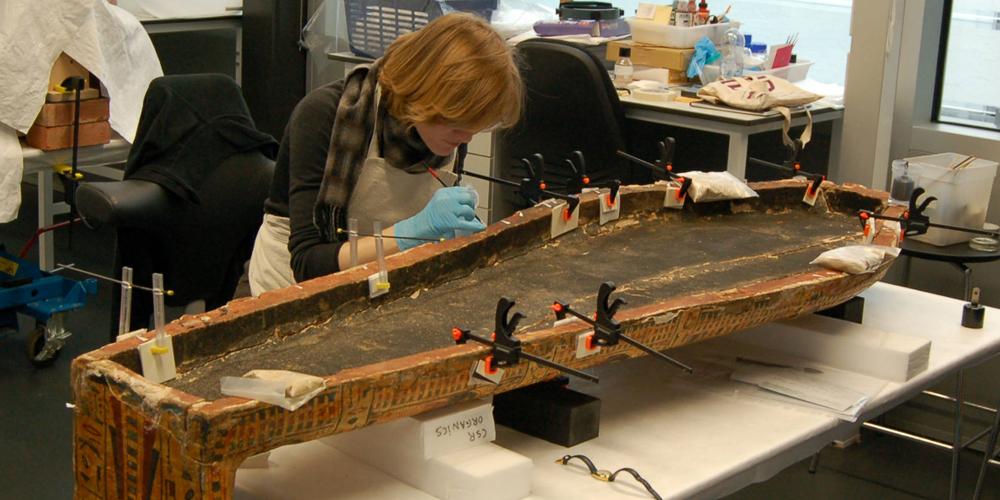 A conservator bending over to work closely on a coffin lid, placed on work table within conservation studio.