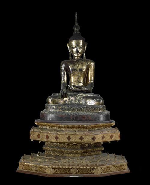 A gilded, lacquered figure of a Buddha in a cross-legged position. The figure sits on a plinth adorned with scrolls of leaves and blossoms.