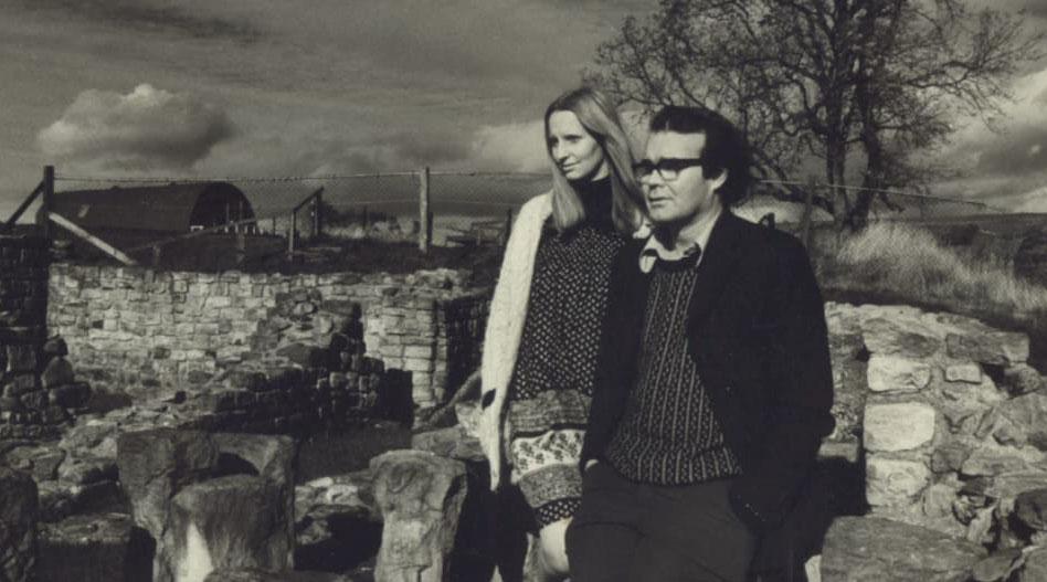 A man in a jumper, blazer and spectacles stands with a blonde-haired woman amongst the ruins of a stone building in a rural landscape