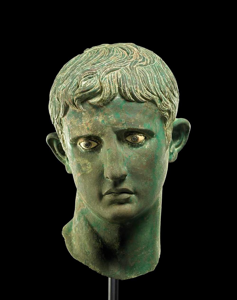 A bronze sculpture of the head of a man with a melancholy expression. The metal is shades of green and bronze