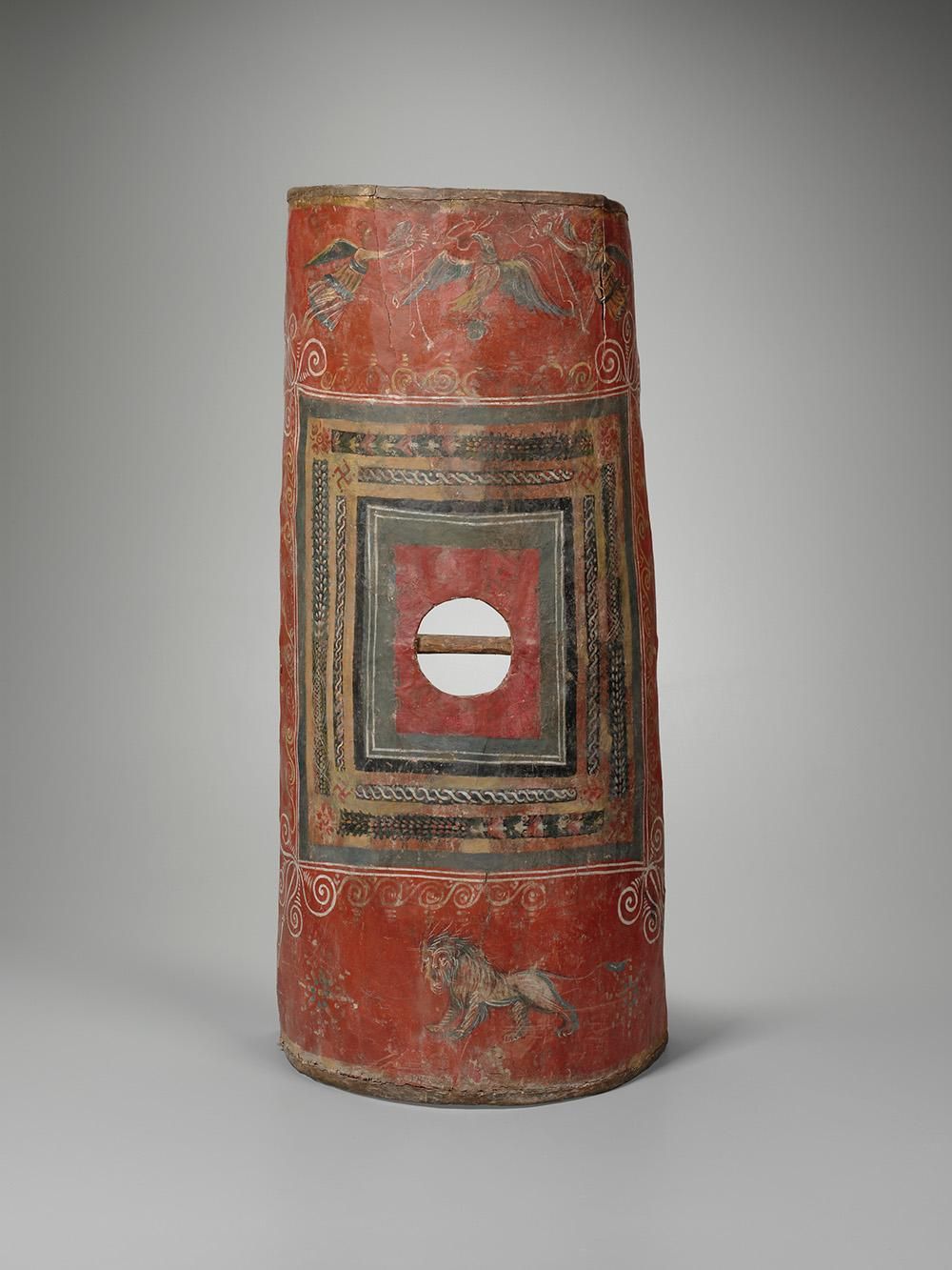 A long soldier's shield made of wood and leather is painted a rich red with motifs of an eagle, a lion and a square geometric pattern