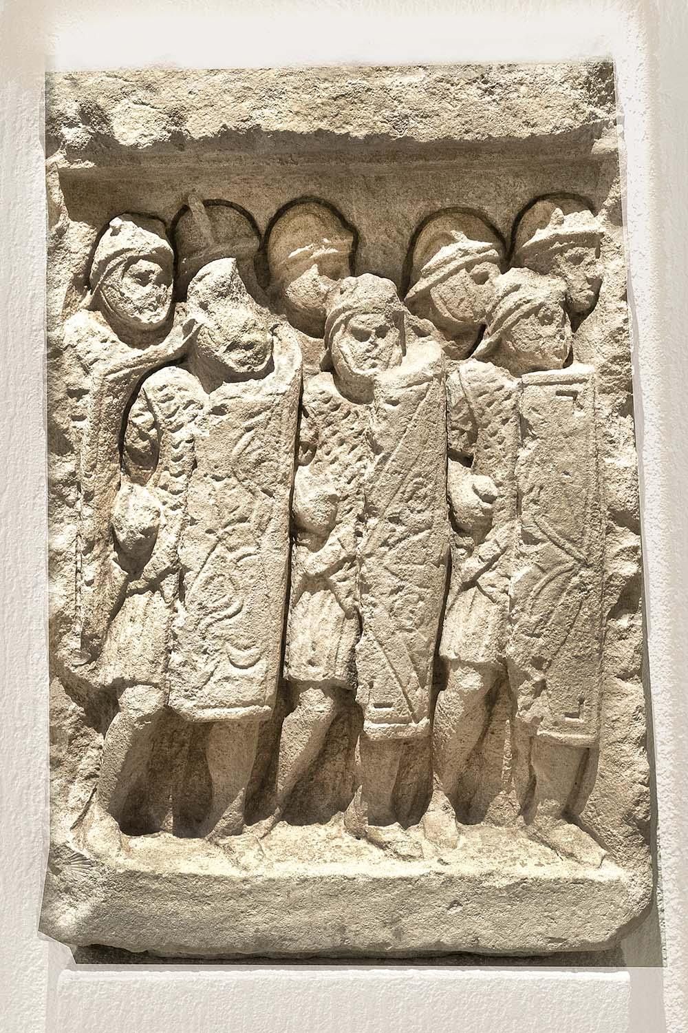  A stone carving shows a group of soldiers tightly packed into a battle formation, wearing helmets and with long shields covering their bodie