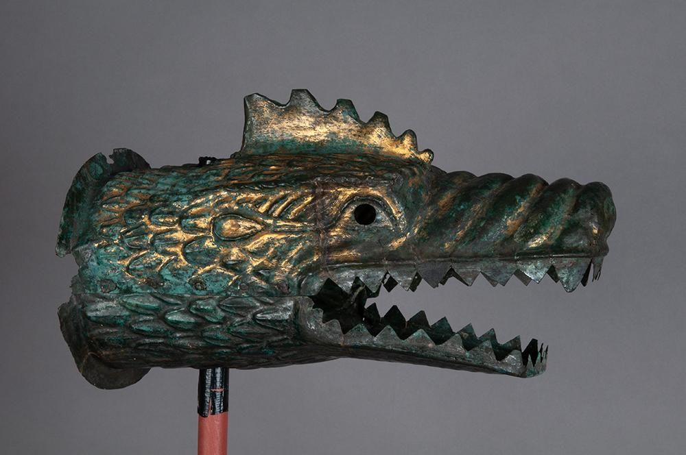 A bronze sculpture of a dragon head with an open mouth and sharp teeth, flecked in shades of golden metal and green oxidised metal