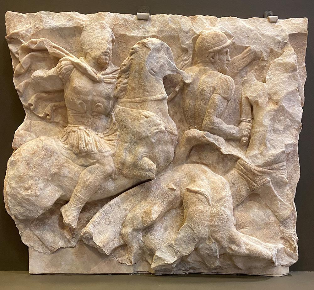 A stone relief of a man with a cloak riding a horse which tramples a person beneath it, and another soldier holding a sword.