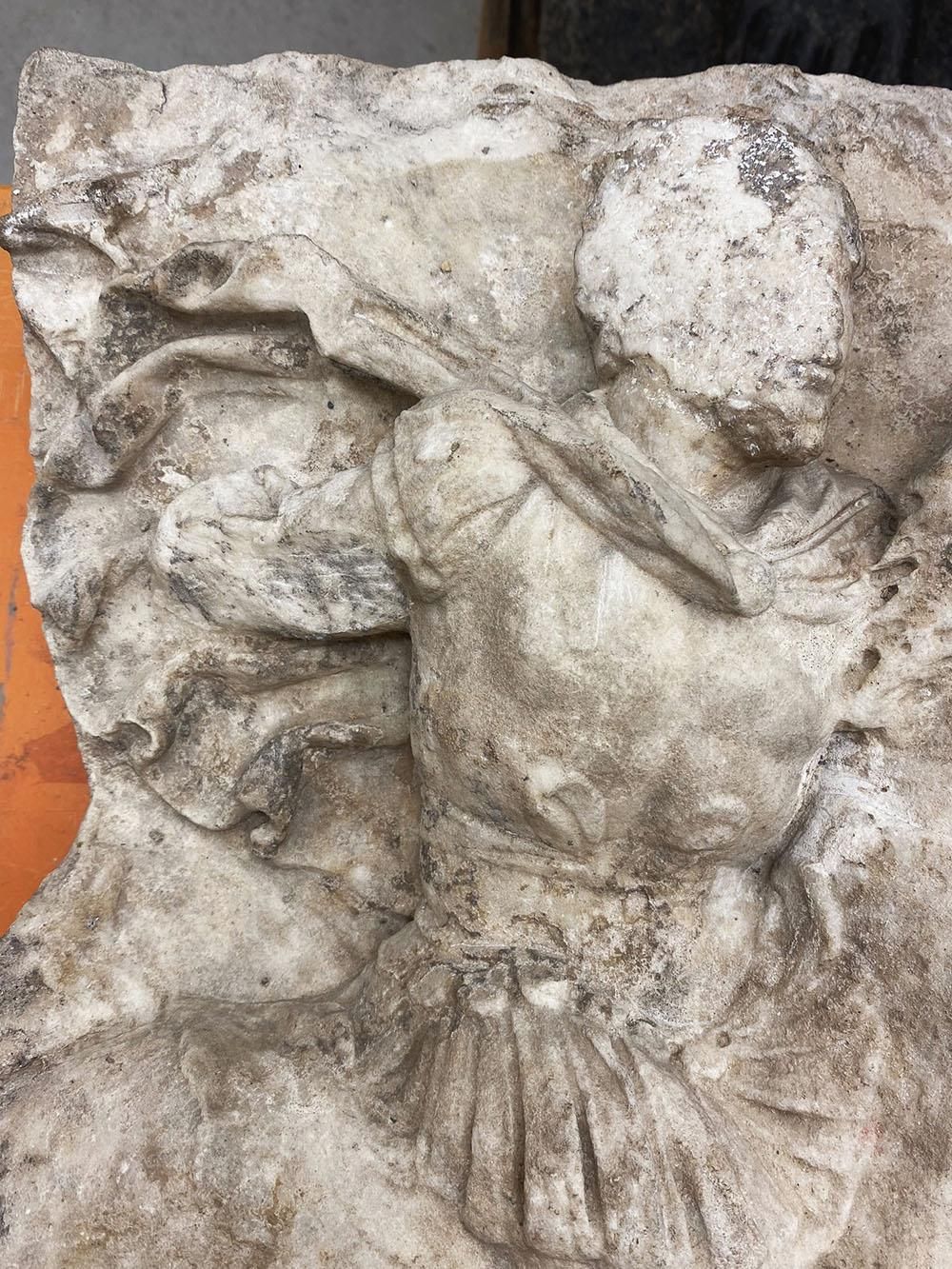 A detail of a stone relief showing a man riding a horse with cloak flowing behind. The face is damaged and missing.