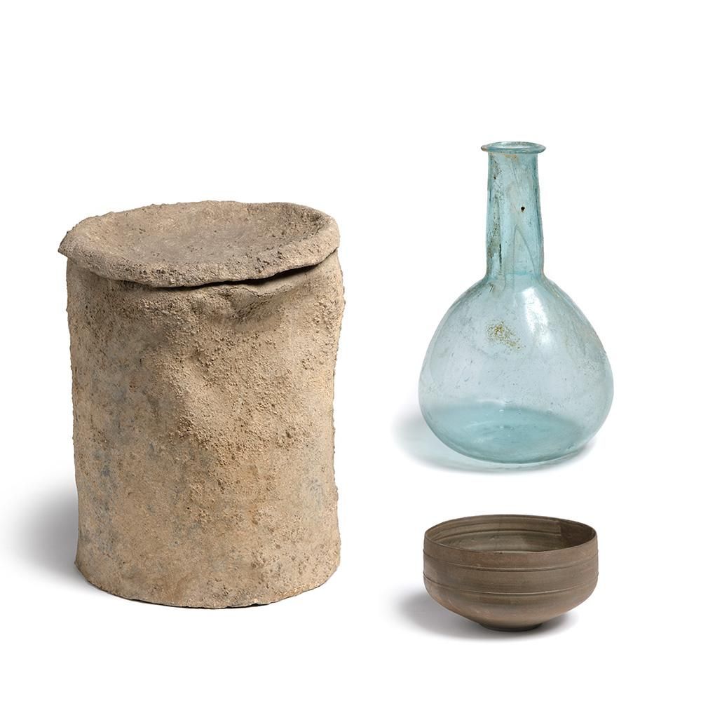 A rough clay cylinder pot with lid, next to a small thrown clay cup, and a hand-blown glass bottle with a spherical base and a long narrow neck. The glass is a pale blue-turquoise with flecks of rust.