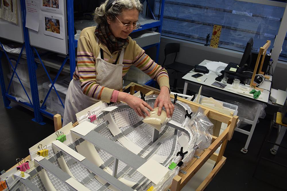 A woman with grey hair and glasses works on a mount for the mummy case