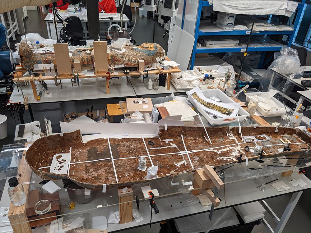 The lid and base of a cartonnage coffin in the conservation lab, supported by structures and with conservation materials surrounding them