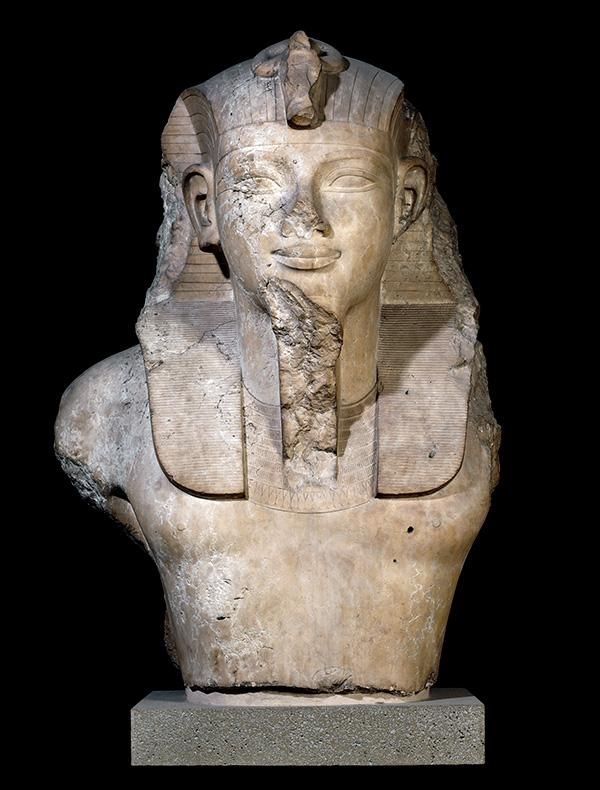 A worn and chipped statue of a Egyptian king, upper body from chest to face visible. The figure is wearing a head-dress and there is damage to the sculpture on his chin, shoulder and headpiece.