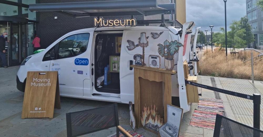 White van parked up with an open door and some furniture resting outside, including a sign with the words "Moving Museum".