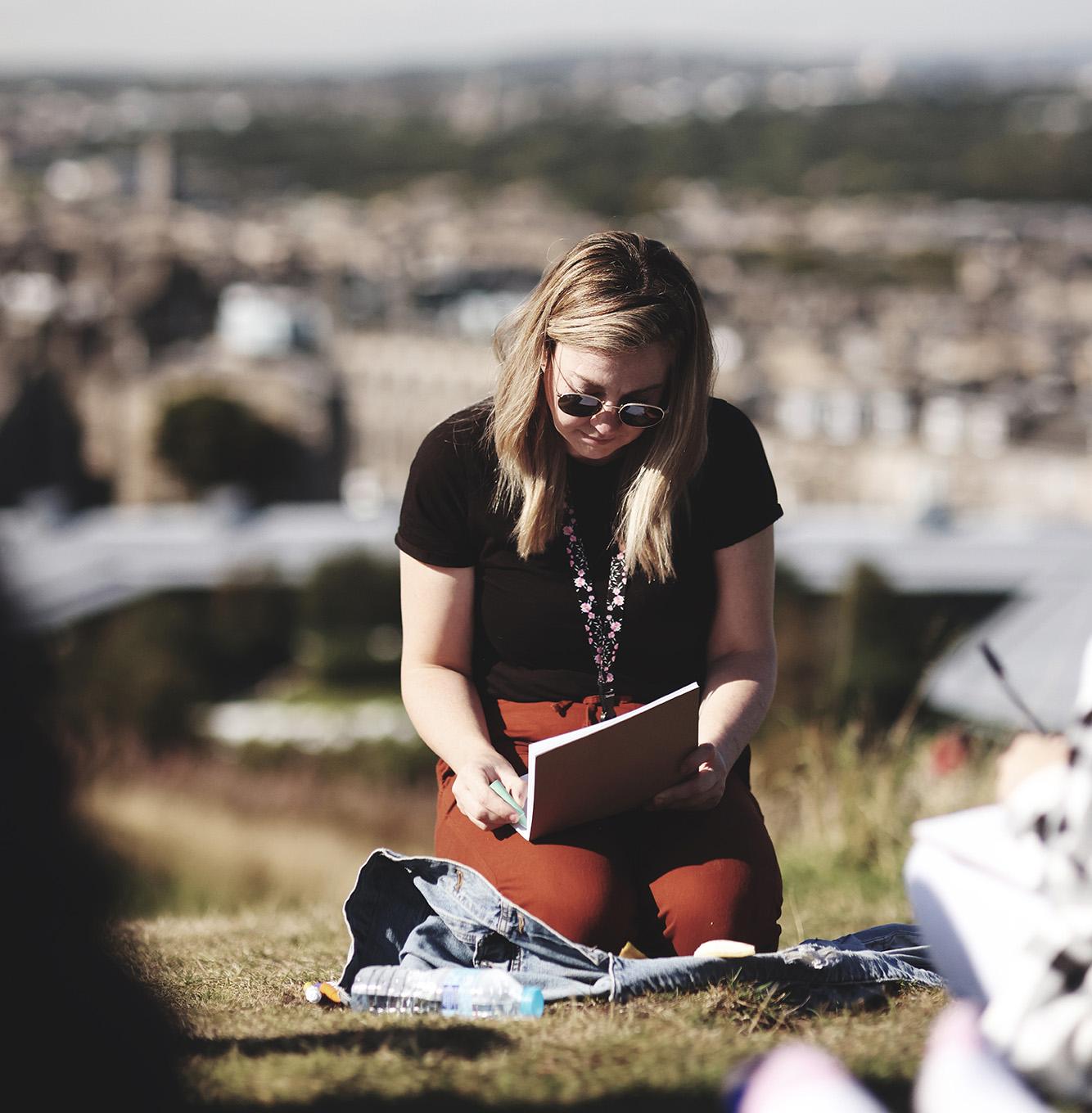 Young person with a sketchbook on a hill with Edinburgh city in the distance.